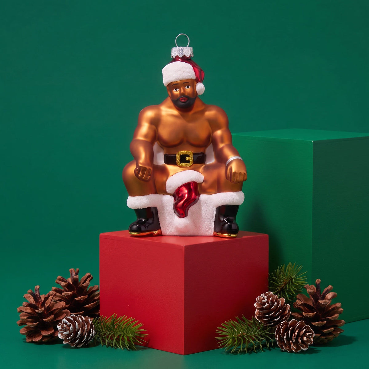 A festive Christmas ornament shaped like a muscular man wearing a Santa hat and Santa pants, seated on a red pedestal against a green background with pinecones and small greenery around it.