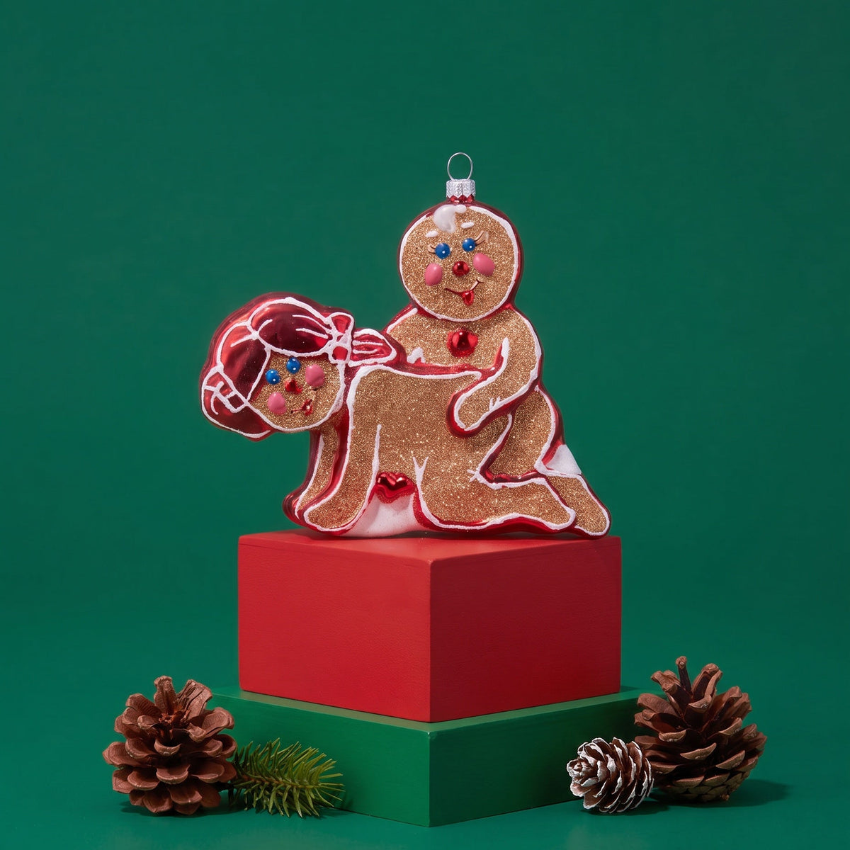 A festive Christmas ornament shaped like two gingerbread figures, one larger figure standing on a red cube and one smaller figure seated beside it, styled with pinecones on a green background.