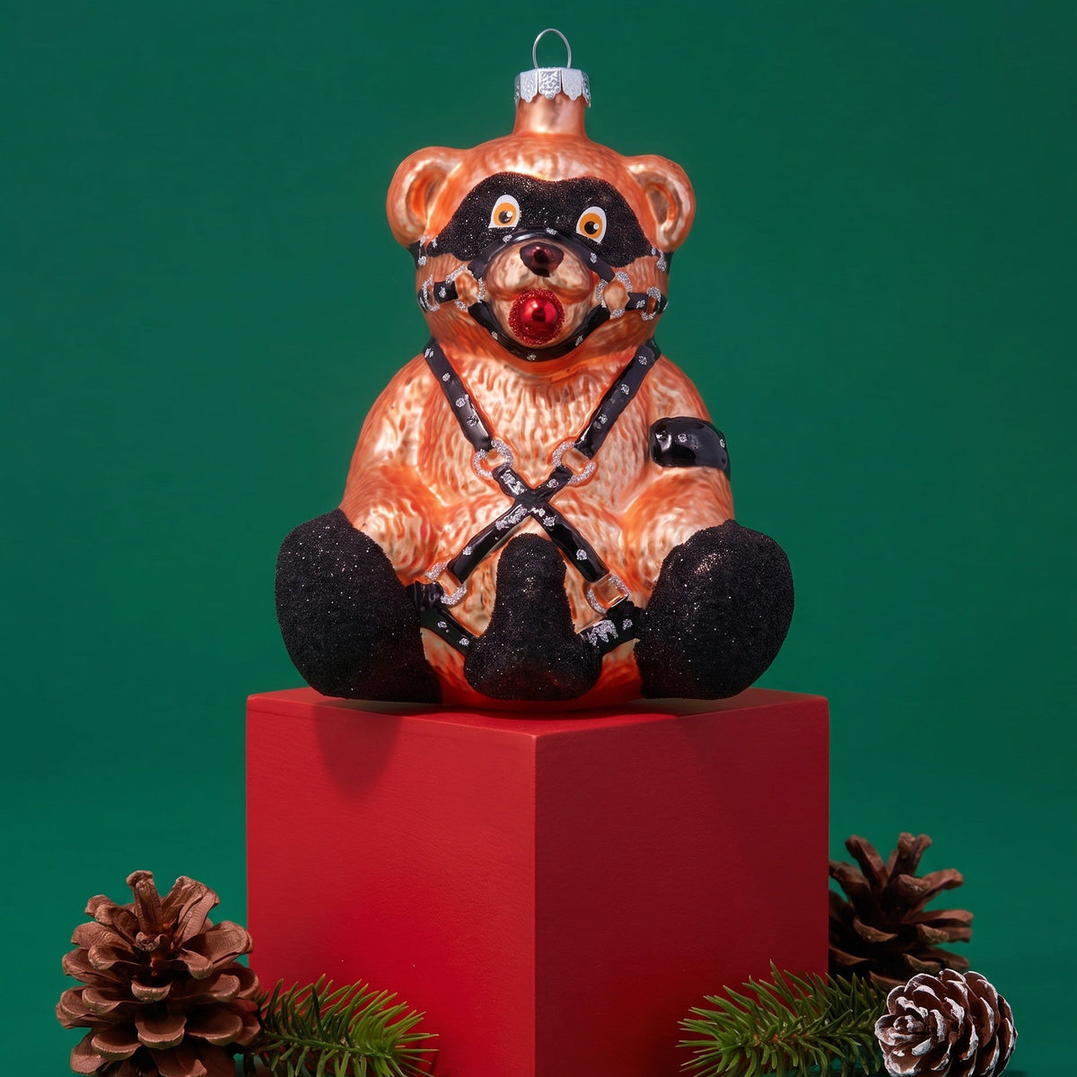 A vintage-style Christmas ornament shaped like a seated bear, with a glossy orange-brown body, black face and paws, and a red nose, displayed on a red pedestal with pinecones against a green background.
