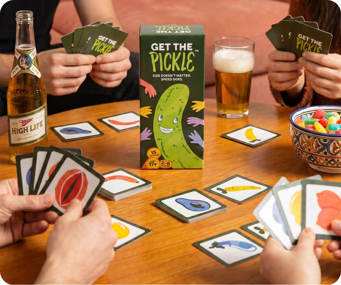 Players holding cards around a wooden table playing Get the Pickle, with the product box in the center, a bottle of Miller High Life beer, a glass of beer, and a bowl of candy.