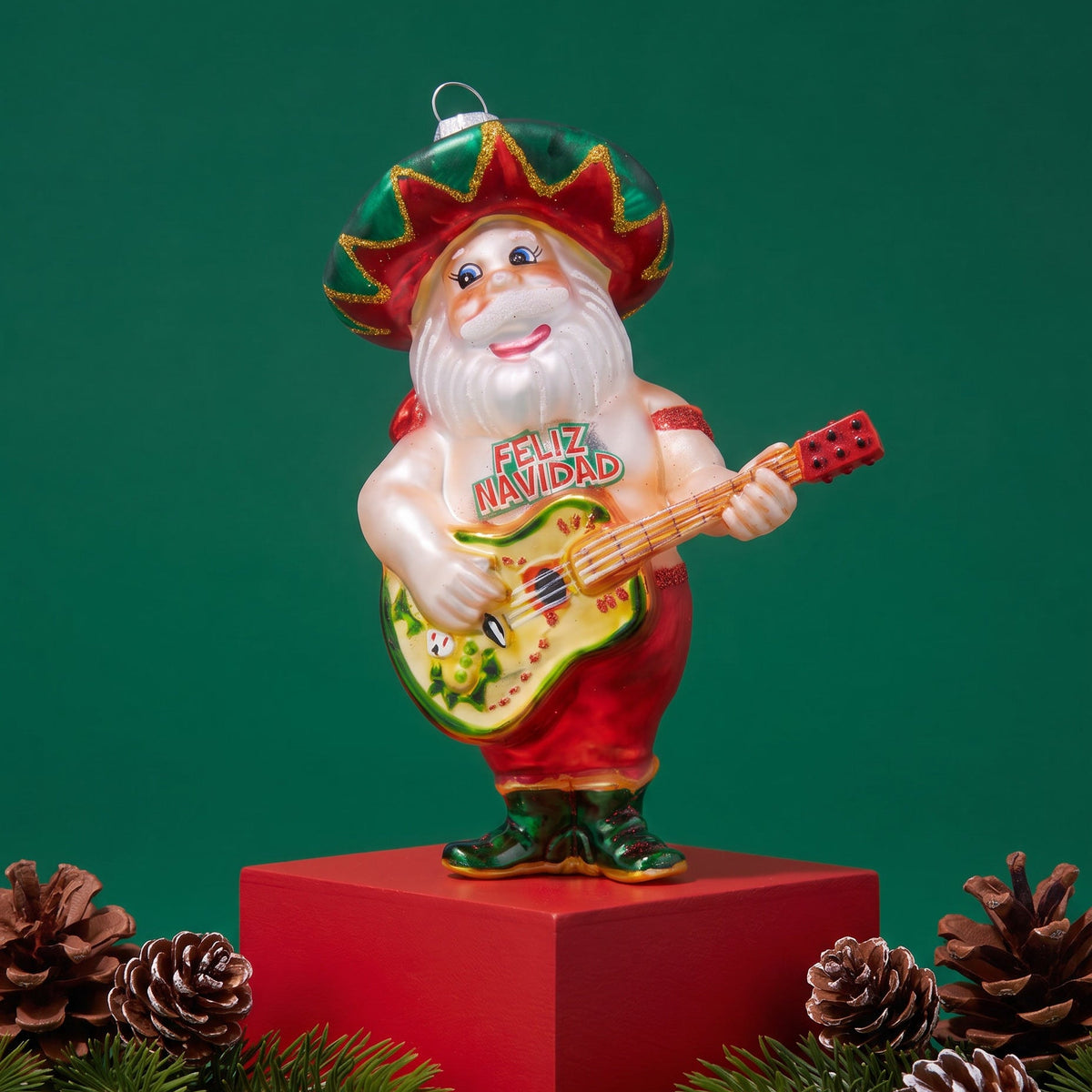 A festive Christmas ornament shaped like a Santa-like figure wearing a green and red sombrero and playing a guitar, displayed on a red pedestal with pine branches and pinecones against a dark green background.