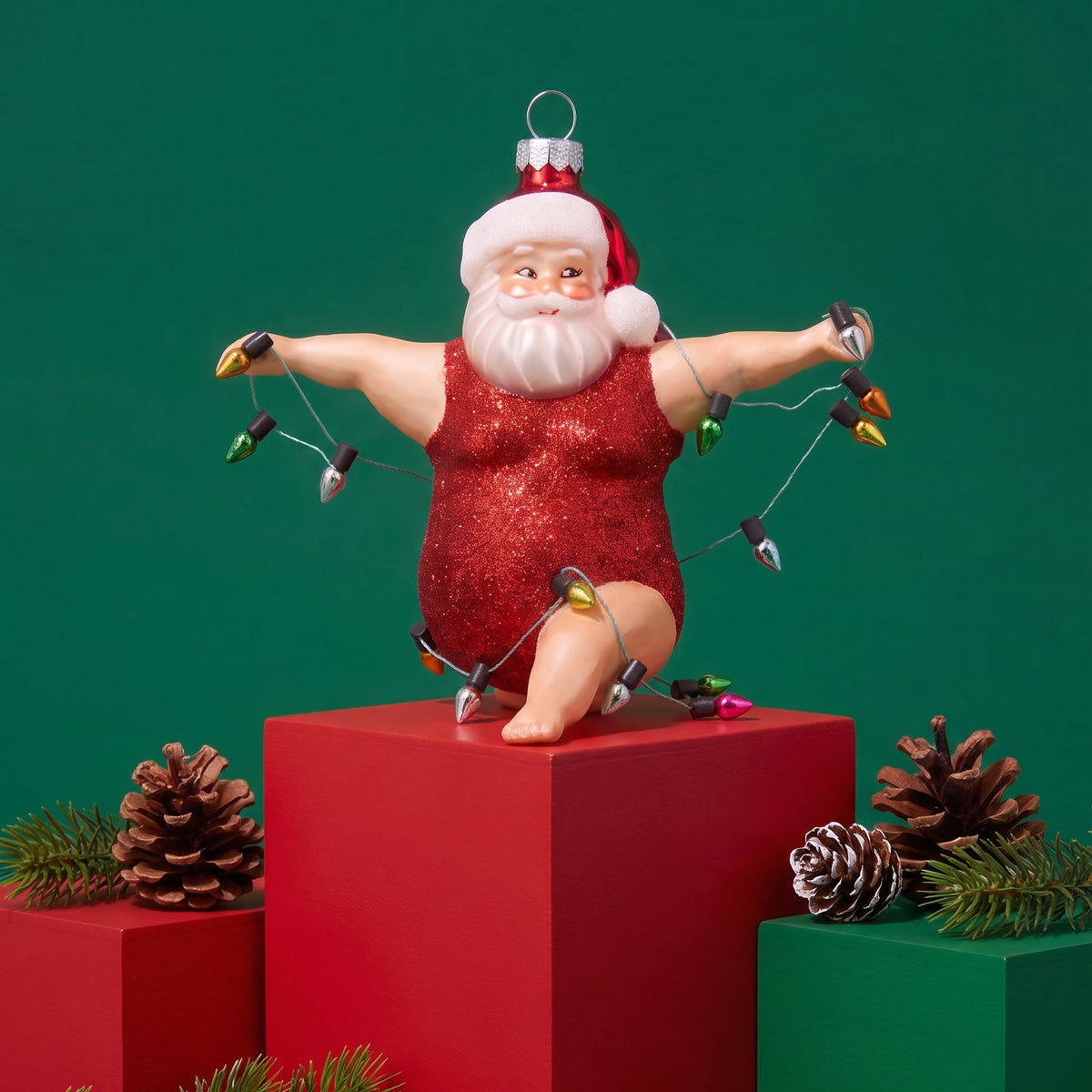 A festive Christmas ornament shaped like a Santa Claus figure in a glittery red romper, posed on red and green pedestals with pinecones and evergreen branches against a solid green background.